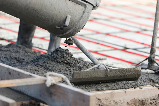 Concrete is being poured from a mixer onto red and grey mesh flooring. A rake spreads the wet concrete, suggesting ongoing construction work.