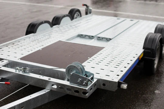A flatbed car trailer with multiple axles, featuring a metallic perforated surface and winch mechanism, is parked on a wet, overcast asphalt area.