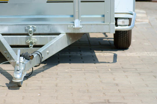 Close-up of a metal trailer hitch on a paved surface, showing a robust, geometric structure with a circular connector. Sunlight casts a shadow.