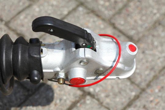 Close-up of a metal trailer hitch with a black handle and red safety cable on a brick-paved surface, suggesting security and durability.
